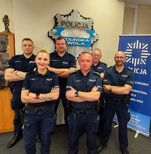Sześciu umundurowanych policjantów stoi pod ścianą, za nimi gwiazda policyjna z napisem Zduńska Wola. Pozują do zdjęcia.