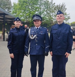 Do zdjęcia pozuje troje policjantów. Po środku stoi w mundurze galowym zastępca komendanta. Po lewej stronie stoi kobieta w mundurze, a po prawej mężczyzna w mundurze.