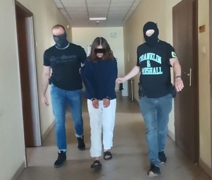 Dwaj policjanci prowadzą kobietę, która ma założone na rekach kajdanki.