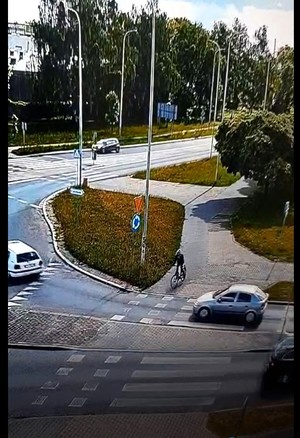 Zdjęcie ulicy w mieście, do przejazdu dojeżdża rowerzystka, a od prawej strony do przejazdu zbliża się samochód.