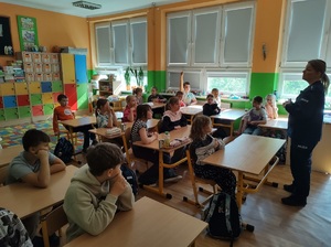 Klasa, uczniowie siedzą w ławkach szkolnych, przed nimi stoi umundurowana policjantka, mówi do dzieci.