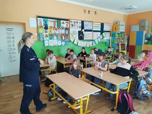Klasa, uczniowie siedzą w ławkach szkolnych, przed nimi stoi umundurowana policjantka, mówi do dzieci.