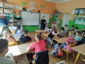 Klasa, uczniowie siedzą w ławkach szkolnych, przed nimi stoi umundurowana policjantka, mówi do dzieci.