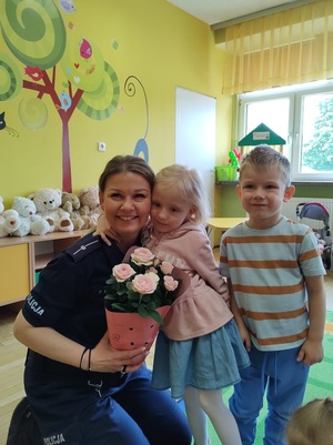 Policjantka pozuje do zdjęcia z kwiatkami w doniczce. Przytula się do niej dziewczynka, obok stoi chłopiec.