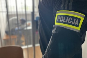 Policjant stoi bokiem, na ramieniu ma opaskę z napisem policja. W tle, za kratkami siedzi mężczyzna.
