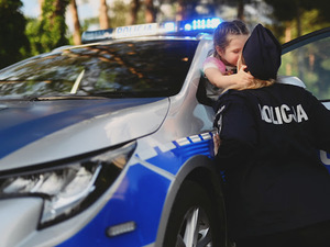 Dziewczynka wychyla się z okna w radiowozie, przed nią jest kobieta ubrana w policyjny mundur. Dziewczynka kobietę przytula i całuje.