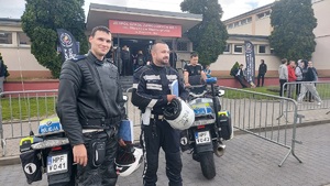 Policjanci w strojach motocyklowych, stoją koło motocykli, przed budynkiem szkoły. W tle widać chodzącą młodzież.