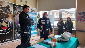 Pomieszczenie wewnątrz szkoły. Dwaj policjanci pozują uśmiechnięci do zdjęcia, w tle widać dwie uczennice.