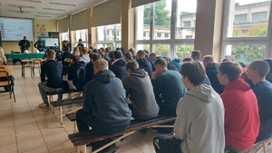 Pomieszczenie w szkole. Tyłem do zdjęcia siedzą uczniowie. W tle widać policjantów, którzy mówią do zebranych.