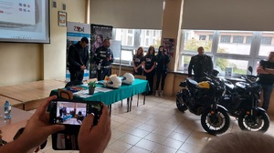 Policjanci stoją przy stole, mówią do zebranych, za nimi pod oknem stoją dwie dziewczyny.