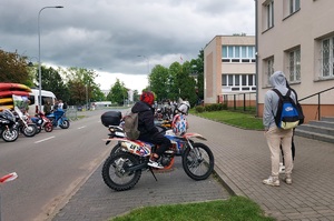 Teren przed szkoła. W dwóch rzędach stoją motocykle. Na jednym z nich siedzi chłopak tyłem do zdjęcia.