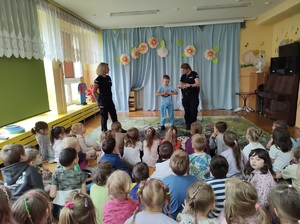 Sala w przedszkolu. Na dywanie siedzą dzieci, przed nimi stoją dwie policjantki i chłopiec.