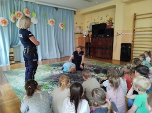 Sala w przedszkolu. Na dywanie siedzą dzieci, przed nimi stoi jedna policjantka, a druga razem z chłopcem siedzi na dywanie. Chłopiec pokazuje jak wygląda bezpieczna pozycja na żółwia w przypadku ataku przez agresywnego psa.