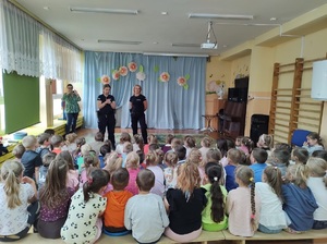 Sala w przedszkolu. Na dywanie siedzą dzieci, przed nimi stoją dwie policjantki, które mówią do dzieci.
