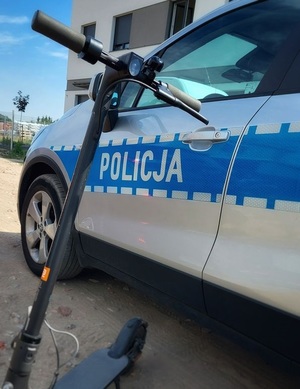 Na zdjęciu hulajnoga elektryczna, a z tyłu za nią stoi radiowóz.