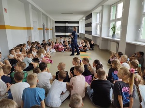 Korytarz w szkole. Dzieci siedzą na podłodze, słuchają co mówi do nich policjantka.