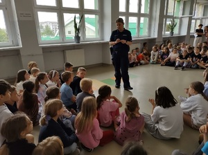 Korytarz w szkole. Dzieci siedzą na podłodze, słuchają co mówi do nich policjantka.