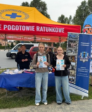Dwie nastolatki stoją, trzymają w rękach ulotki dotyczące bezpiecznych wakacji, za nimi pod namiotem stoi policjantka.