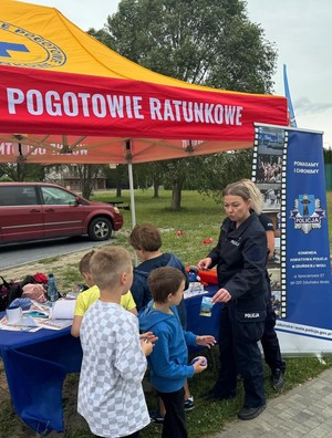 Dzieci i policjantki stoją przy stoisku policyjnym. Policjantka daje dzieciom ulotki o bezpieczeństwie.