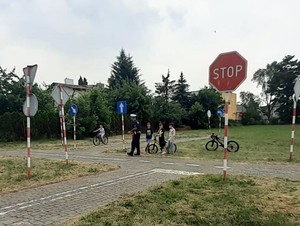 Policjant stoi z uczniami, obserwują jak chłopak na rowerze jedzie torem gdzie są znaki drogowe imitujące ulicę.