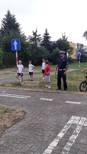 Policjant stoi z uczniami, obserwują jak chłopak na rowerze jedzie torem gdzie są znaki drogowe imitujące ulicę.