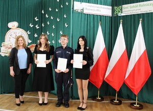 Troje uczniów pozuje do zdjęcia z książkami i świadectwami. Obok nich stoi kobieta.