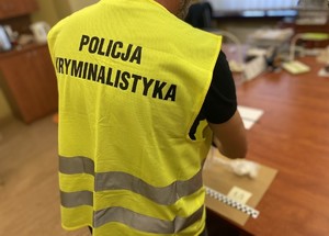 Tyłem stoi policjant, ma założoną odblaskową kamizelkę z napisem: Policja kryminalistyka