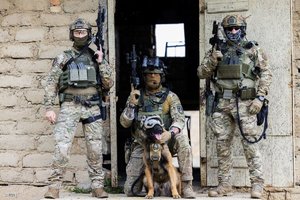 Trzej policjanci z psem, ubrani w mundur służb kontrterrorystycznych, pozują do zdjęcia na tle budynku.