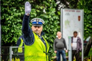 Policjant ruchu drogowego jest na ulicy, kieruje ruchem, ma podniesioną do góry rękę, w ustach ma gwizdek.