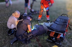 Na trawniku leży na boku osoba, pochyla się nad nią policjant, dwóch ratowników medycznych i kobieta.