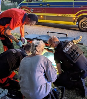 Na trawniku leży na boku osoba, pochyla się nad nią policjant, dwóch ratowników medycznych i kobieta. W tle widać nosze i karetkę pogotowia.