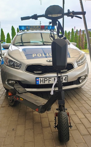 Hulajnoga elektryczna, za nią stoi radiowóz policyjny.