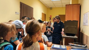 W wąskim korytarzu stoją dzieci, słuchają policjantki, która stoi obok i mówi do nich.