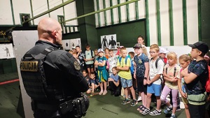 Dzieci są na strzelnicy. Po lewej stronie stoi policjant - instruktor wyszkolenia strzeleckiego, mówi do dzieci.