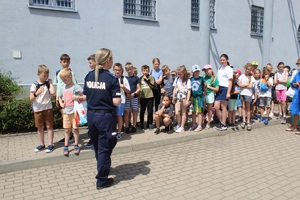 Teren na zewnątrz komendy, dzieci stoją na chodniku, przed nimi stoi policjantka, mówi do nich.