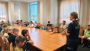 Pomieszczenie wewnątrz komendy. Dzieci siedzą na krzesłach za stołem, przed nimi stoi policjantka i mówi do nich.