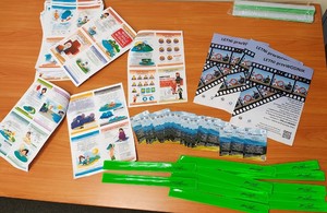 Na stole są przygotowane dla dzieci opaski odblaskowe, ulotki i plakaty dotyczące bezpieczeństwa podczas wakacji.