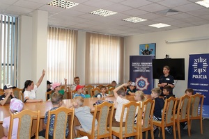 Aula w komendzie. Dzieci siedzą na krzesełkach przy stole, w szczycie stołu stoi policjantka i mówi do dzieci.