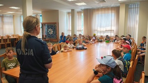Policjantka mówi do dzieci, siedzących na krzesłach przy długim stole.