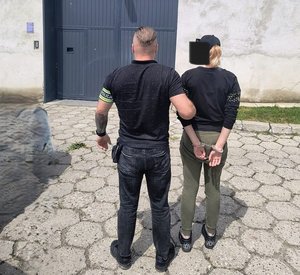 Po prawej stronie stoi zatrzymana, ma założone kajdanki, po jej lewej stronie stoi ubrany po cywilnemu policjant, trzyma ją za ramię, przed nimi jest brama więzienia.