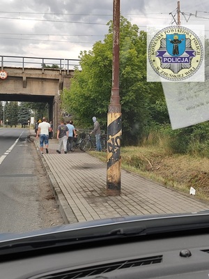 po lewej stronie ulica, po prawej chodnik, na chodniku stoi słup latarni, w tle widać pieszych, przechodzących pod wiaduktem. U góry po prawej stronie logo zduńskowolskiej komendy.