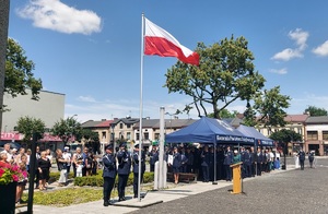 Na maszcie powiewa flaga, pod maszem stoi trzech policjantów. Dalej jest namiot, pod którym stoją goście i policjanci.