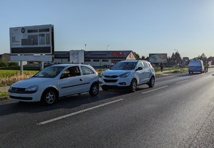 Droga, wzdłuż niej po lewej stronie stoją dwa samochody jeden za drugim, w tle widać sklep spożywczy oraz radiowóz.