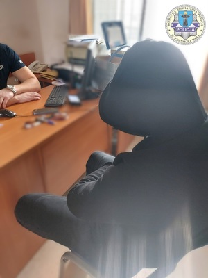 Zatrzymany siedzi tyłem do zdjęcia, ma założony kaptur na głowę, siedzi przy biurku, na wprost niego siedzi umundurowany policjant.
