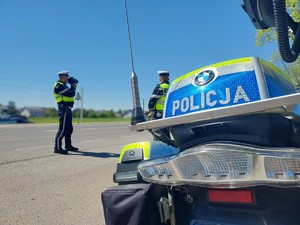 Przy drodze stoi zaparkowany motocykl służbowy, w tle stoją dwaj umundurowani policjanci, jeden z nich patrzy na drogę przez urządzenie, które mierzy prędkość jadących pojazdów.