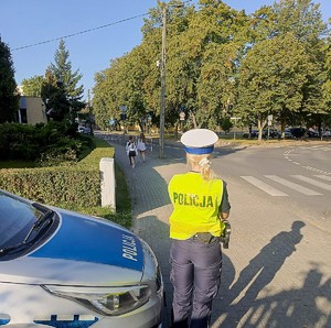 Policjantka stoi przy radiowozie, w pobliżu przejścia dla pieszych. Patrzy, jak chodnikiem idą dzieci.