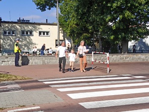 Policjant stoi pod szkołą przy przejściu dla pieszych. Do przejścia dochodzi dziecko, trzymając dwie dorosłe osoby za ręce.