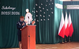 Przy mównicy stoi policjantka, mówi do mikrofonu. Po prawej stronie na krzesełkach siedzą uczniowie, obok stoją trzy flagi państwowe. Za kobietą w tle jest ścianka na której jest napis Rozpoczęcie Roku Szkolnego 2025/2026.