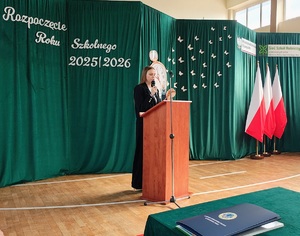 Przy mównicy stoi uczennica, mówi do mikrofonu. Po prawej stronie stoją trzy flagi państwowe. Za kobietą w tle jest ścianka na której jest napis Rozpoczęcie Roku Szkolnego 2025/2026.