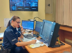 Dyżurna siedzi przed monitorami, odbiera telefon, zapisuje coś na kartce.
